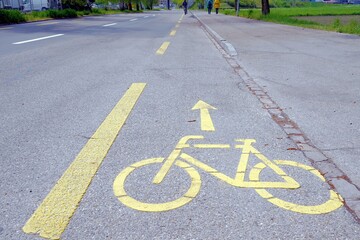 Obraz premium Bicycle lane with yellow marking on the ground in Switzerland. On the right, there is sidewalk for pedestrians. In the background are silhouettes of pedestrians and one person on bike.