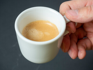 a man's hand holds a white cup of freshly brewed espresso on a dark background. Brown foam. An invigorating drink. top view