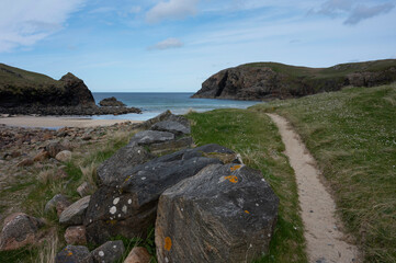 Sandy path to Dail Beag beach on Isle of Lewis, Outer Hebrides, Scotland. Rocks and grass at side of path, leading to beach, sea and dramatic cliffs. Blue sky and clouds.