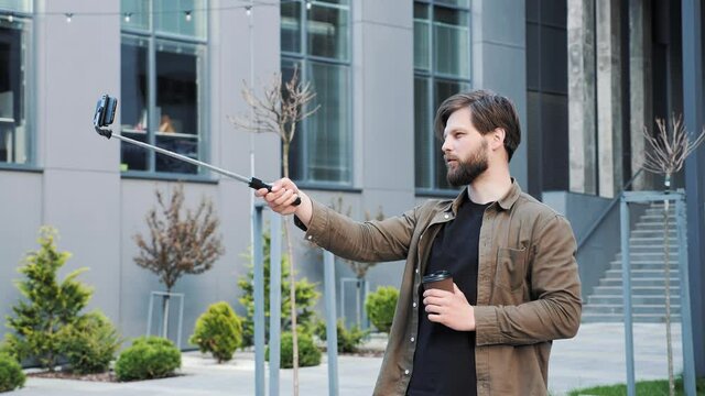 Young bearded man holding smartphone on the tripod shooting his blog standing outside near modern buildings on the background. Blogging shoot video using mobile phone for social network content.