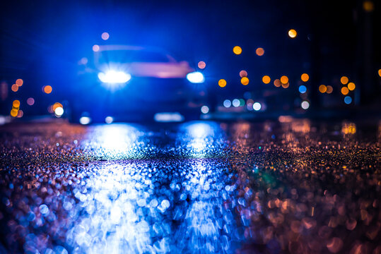 Rainy Night In The Big City, Blinding Headlights Of An Approaching Car. View From The Level Of Asphalt