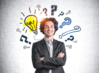 Portrait of handsome hispanic businessman in suit with crossed arms pose and wondering about career opportunities. Lightbulb and question marks drawn on concrete wall behind. Searching for answer.