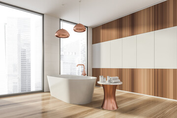 Fototapeta na wymiar Modern design bathroom interior with white oval bathtub, bronze faucet, lamp. Small table with towel and cosmetics. Panoramic window with skyscrapers city view. Wood materials.