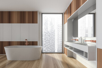 Modern design bathroom interior with white oval bathtub, double sink countertop, bronze faucets. Panoramic window with skyscrapers city view. Wooden and white ceramic tile on wall.