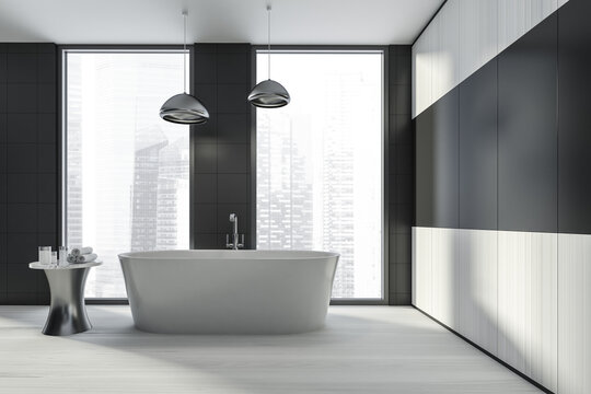 Modern design bathroom interior with white oval bathtub, silver faucet, lamp. Small table with towel and cosmetics. Panoramic window with skyscrapers city view. Wood materials.