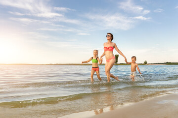 Young adult attractive slim sporty mother enjoy having fun running water by lake or sea sand breach with two cute little siblings against blue sky on summer day. Summertime family vacation concept
