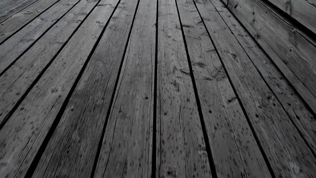 Bouncing walk on wooden quay. Coarse wide planks that are fastened with large nails. You can see the water between the planks in some places. Raindrops have created dark spots on the quay.