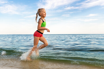 Cute adorable sporty caucasian blond school girl enjoy having fun running water by lake or sea sand beach splashing against blue sky on summer day. Summertime children healthy vacation concept