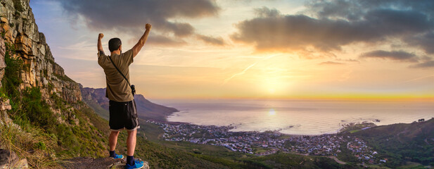 Victory and success after a hike up Table Mountain at sunset with vibrant orange sky - looking out over the bay - Great outdoors adventure and travel holiday destination, Cape Town, South Africa - obrazy, fototapety, plakaty
