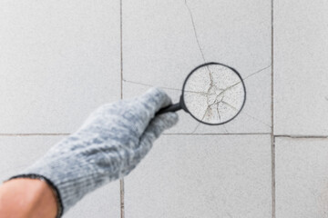 The hand of an industrial worker in a construction glove with a magnifying glass examines the damaged cracked gray tile background. Renovation concept