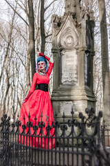 Beautiful gothic model at the old monument. Bright red vintage dress and unusual makeup. Tall spring trees and a wrought-iron fence.