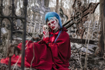 Sensual gothic beauty in the cemetery. Beautiful model with a skull sitting at the cross. Unusual red makeup and cape.