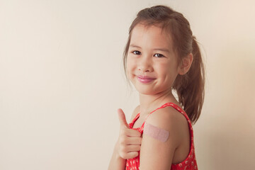 Happy mixed Asian girl  giving thumb up and showing her arm with bandage after got vaccinated or  inoculation, child immunization - obrazy, fototapety, plakaty