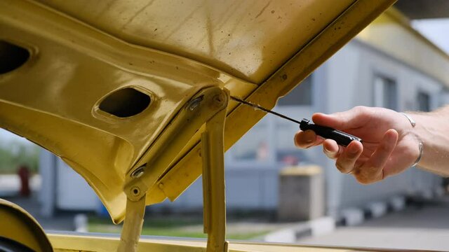 male man mechanic applies lubricant to hood hinges of old beige retro car with screwdriver. Repair on the road.