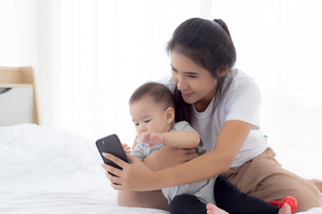 Fototapeta na wymiar Young asian mother and little baby girl or newborn selfie with smart phone on bed in bedroom, happiness mom and daughter using phone video call at home, two people, family and communication concept.