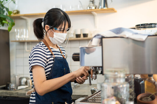 Asian female barista wearing face mask and shield, using coffee maker.