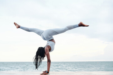 Side view of athletic young woman practicing yoga, doing handstand with splits pose on beach