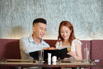 Joyful young couple sitting at restaurant table, reading menu and choosing food for dinner