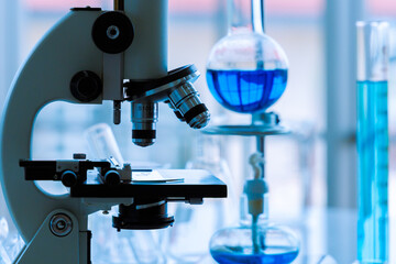 Closeup shot of microscope with scientific sample on plate under lens in front of blurred volumetric and Erlenmeyer flask measuring cylinder glass test tube and laboratory glassware in background