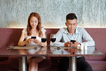 Boyfriend and girlfriend drinking wine, waiting for food and checking social media on smartphones