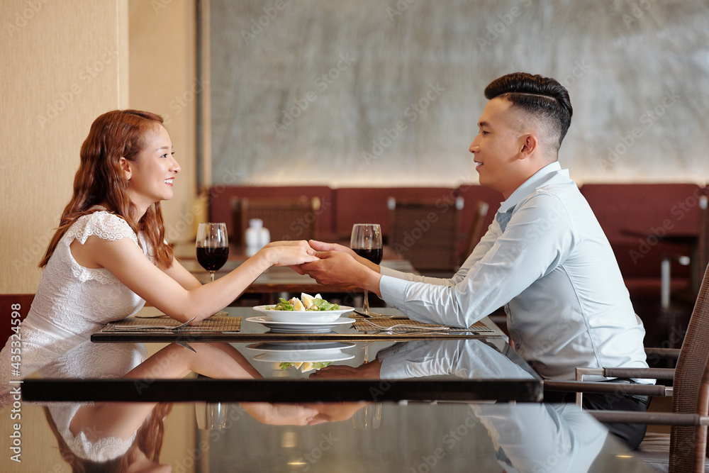 Wall mural Happy young couple in love sitting at restaurant table, holding hands and looking at eyes of each other