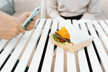 Obraz premium Street fast food. Female hands holding a mobile phone and taking a photo of food, close-up. Photographing a burger on the street, cropped image.
