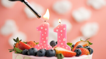 Birthday cake number 11, pink candle on beautiful cake with berries and lighter with fire against background of white clouds and pink sky. Close-up