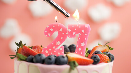 Birthday cake number 27, pink candle on beautiful cake with berries and lighter with fire against background of white clouds and pink sky. Close-up