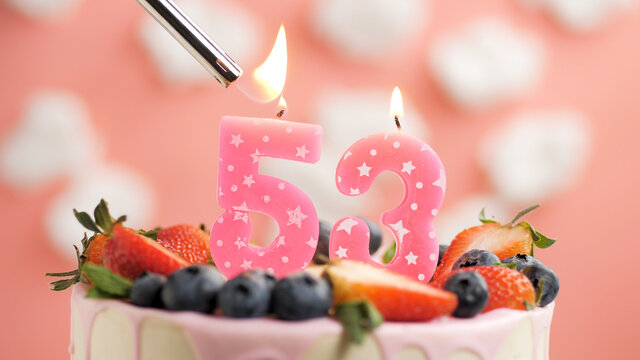 Birthday cake number 53, pink candle on beautiful cake with berries and lighter with fire against background of white clouds and pink sky. Close-up