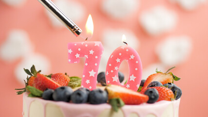 Birthday cake number 70, pink candle on beautiful cake with berries and lighter with fire against background of white clouds and pink sky. Close-up