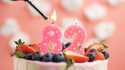 Birthday cake number 82, pink candle on beautiful cake with berries and lighter with fire against background of white clouds and pink sky. Close-up