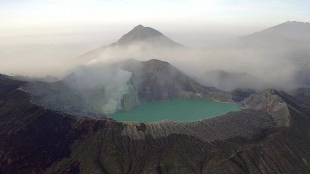 Aerial Circling The Rim Of A Steaming Volcano Caldera, With Jagged Rocks, A Turquoise Lake, And Soaring Foggy Mountain Peaks In The Background - East Java, Indonesia