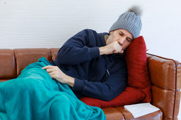 Caucasian sick unwell bearded man in wool jacket knitted hat lay down on leather sofa and pillow in...