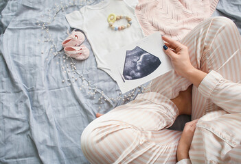 Pregnant woman with sonogram image and baby accessories on bed, closeup