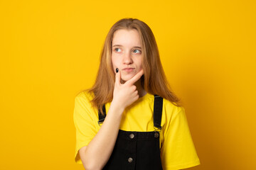 Pleasant looking american teen girl holds chin and looks pensive away, thinks about great plan, has new ideas in mind, wears bright yellow t shirt.