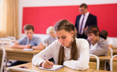 Fototapeta na wymiar Portrait of focused teenage schoolgirl writing lectures in workbooks in classroom during lesson