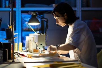 Creating new fashionable collection and garment industry concept. Young female clothes designer sew on sewing machine sitting at workplace in fashion workshop. Small business owner or tailoring hobby