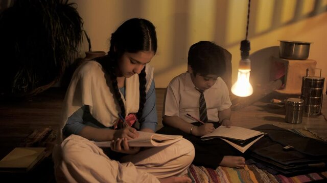 Young boy and girl (Sibling) sitting on the ground and doing homework - Indian village . Medium shot of two small children in school uniform studying under the light of an electric bulb - Rural hom...