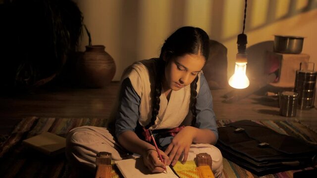 Village student in a school uniform completing homework in a dark village home . Medium shot of young daughter studying alone while sitting near the kitchen area of her house - girl education 