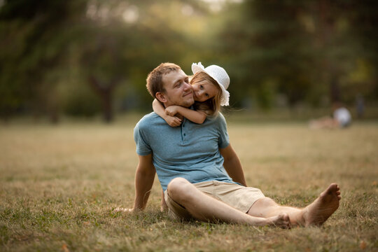Dad and daughter in summer park sit on the grass. Girl hugs and kisses the dad, father smiles and happy. Family leisure concept, nature walk, family care and values, fatherhood, fathers day