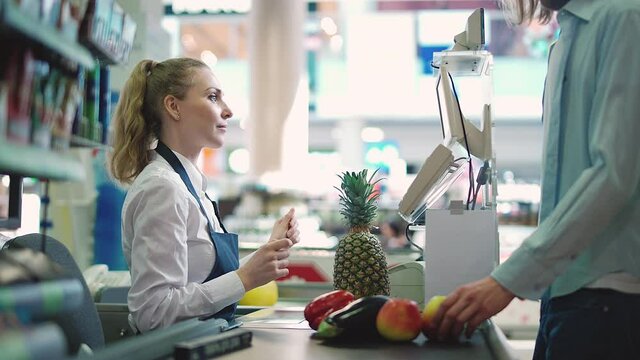 Buying vegetables and fruits in a supermarket, man is pays at the checkout of the grocery store, young woman cashier at work in an organic store.