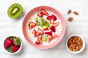 oatmeal porridge with slices of kiwi, strawberries, almonds in pink bowl, spoon on white tablecloth Top view Flat lay Vegan food concept Healthy breakfast