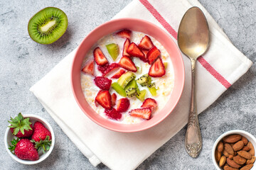 oatmeal porridge with slices of kiwi, strawberries, almonds in pink bowl, spoon, napkin with red stripes on concrete background	