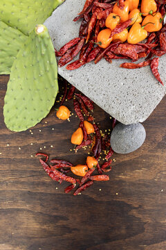 Aerial view of prehispanic foods from Mexico such as metate, chili peppers, prickly cactus, habanero peppers and chile de arbol surrounded by chili seeds on dark wood