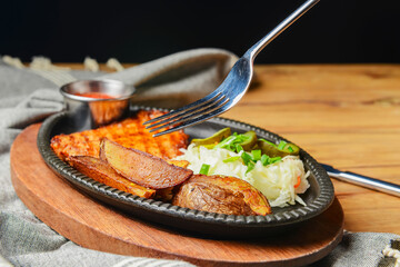 Juicy grilled pork steak with baked potatoes, cut pickles and tomato sauce served in a tray over rustic wooden table.
