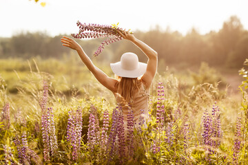 A young girl in a light dress and a hat with a bouquet in her hands on a lupine field, looking at...