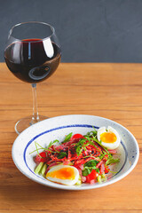Tuna salad with vegetables, boiled eggs, cherry tomatoes and spices and one glass of red wine over wooden background.