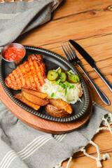 Juicy grilled pork steak with baked potatoes, cut pickles and tomato sauce served in a tray over rustic wooden table.