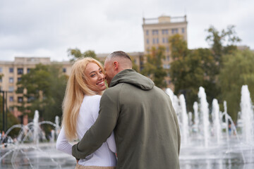 Romantic walk. Young couple in love embracing near fountain in urban park, man whispering compliments to beautiful girlfriend