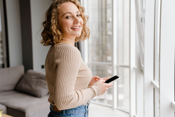Young blonde woman smiling while using mobile phone at home